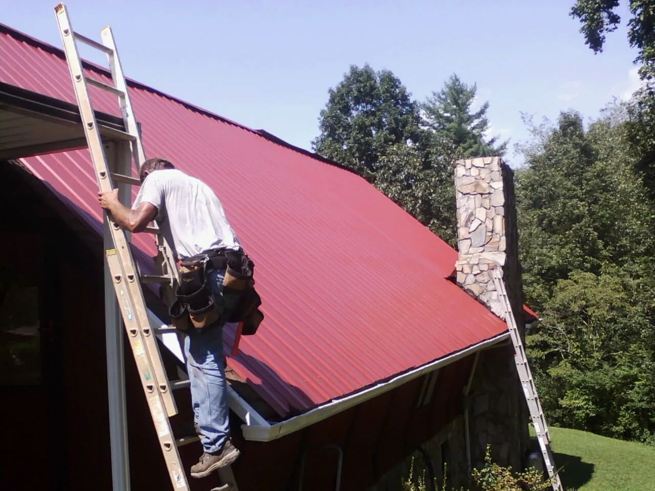 Roofer on a ladder beside a red metal roof and stone chimney — Soffit & Fascia Repair by Roofing Brothers in Hayes